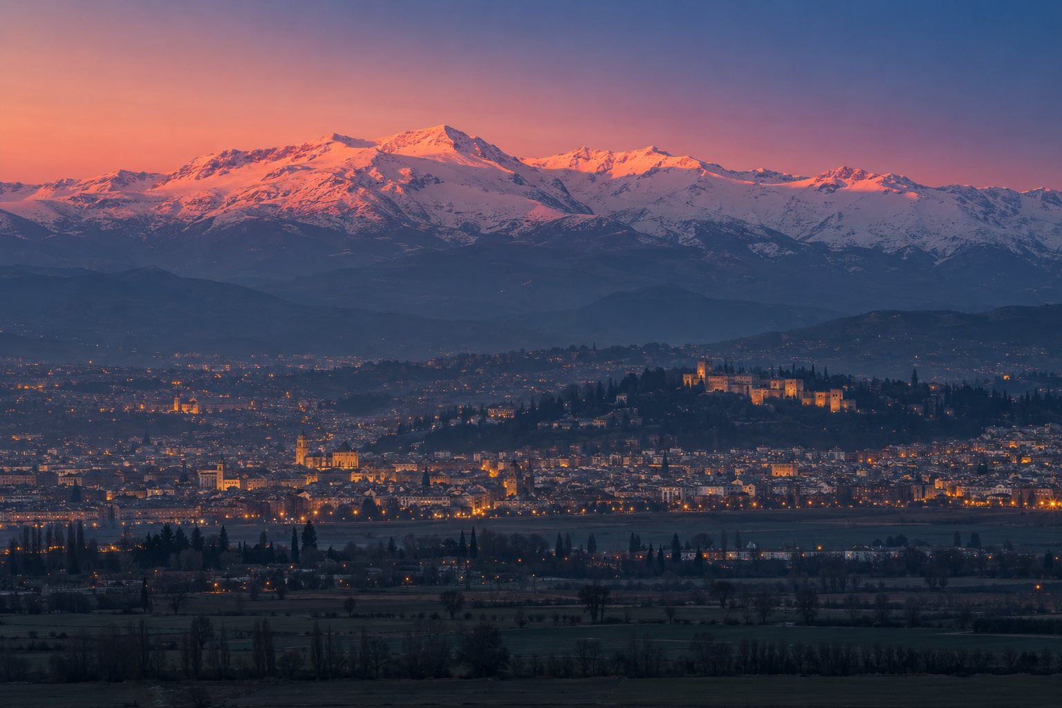Sierra Nevada nevada teñida de rosa al atardecer, con las luces de Granada encendiéndose en la llanura y la Alhambra a lo lejos